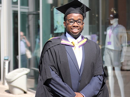 A young black man with glasses and a short beard, wearing a blue suit under black UoM graduation robes and a mortar board. 