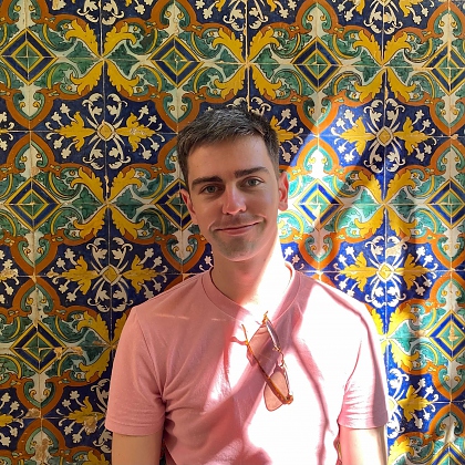 Photo of a young man with short brown hair. He is wearing a pink t-shirt with sunglasses hanging from the collar. He is stood in front of a green, yellow and blue tiled wall. 