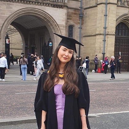 A young woman with long brown hair wearing a light purple dress, black graduation gown and black graduation cap. She is stood in front of the University of Manchester arch on Oxford Road. 