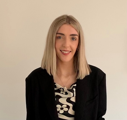 A photo of a smiling young woman with shoulder length blonde hair. She is wearing a black and white blouse and a black blazer. 