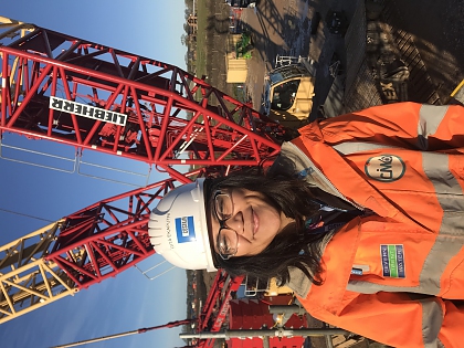 A photo of a young woman with brown wavy hair, wearing a white hard hat and orange hi vis jacket. There is a large red crane in the background.