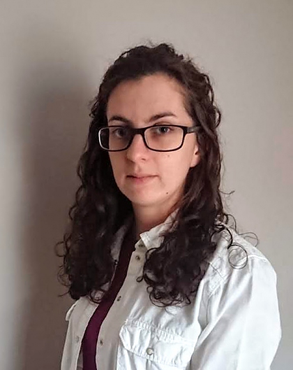 A young woman standing side-on, with dark brown curly hair and black glasses. She is wearing a white shirt. The background is plain. 