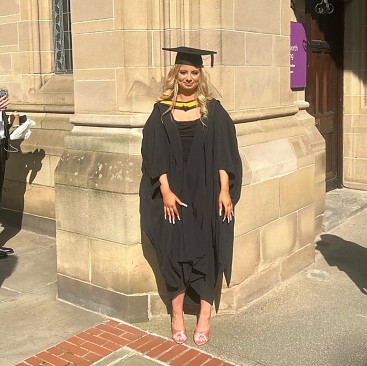 A young woman with blonde hair wearing the black UoM graduation robes and hat, with yellow collar