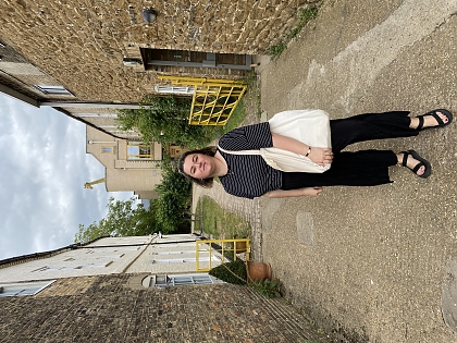 A young woman stood between buildings, with a yellow gate and bushes in the background. She has a large white tote bag and is wearing a striped black t-shirt, black trousers and black sandals. She has short brown hair. 