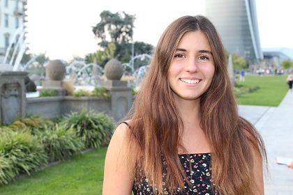 A young woman with long brown hair. She is smiling and is wearing a black top with a small flower pattern. There are gardens behind her. 