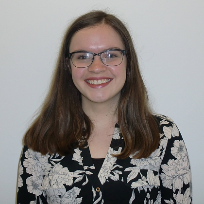 A smiling young woman with long brown hair and black glasses. She is wearing a black and white floral top and is stood in front of a white wall. 