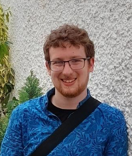 A young man with curly red-brown hair and a beard, wearing glasses and a bright blue top. 