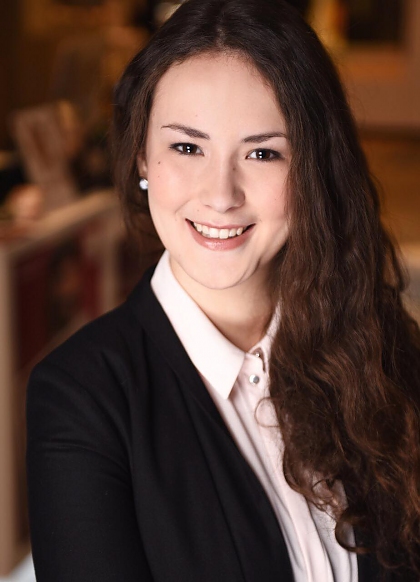 A smiling young woman with long, dark brown, wavy hair. She is wearing a white blouse and black jacket. 