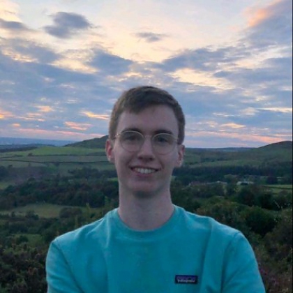 Photo of Aidan wearing glasses and a blue t-shirt with a sunset behind him