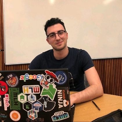 Photo of Enrico wearing a black t-shirt and glasses, sitting in front of a laptop covered in stickers.