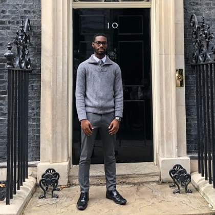 Photo of James in a grey jumper, shirt and tie, standing in front of Number 10, Downing Street.