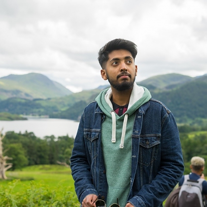 Photo of Shamveel in a green hoodie and denim jacket with lakes and hills behind him