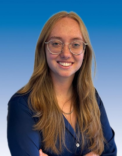 A photo of Katie against a blue blackground. Katie has long blonde hair, glasses and is wearing a blue shirt.